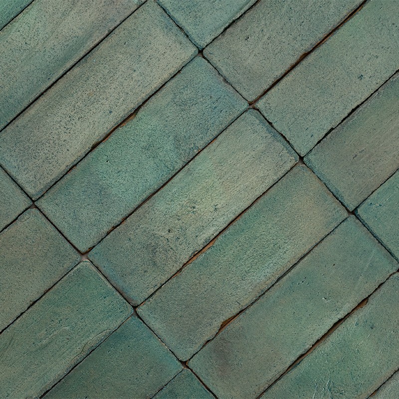 Carrelage rectangulaire turquoise posé en diagonale, joints fins et usure visible, patine verdâtre, fissures et bords brunis.