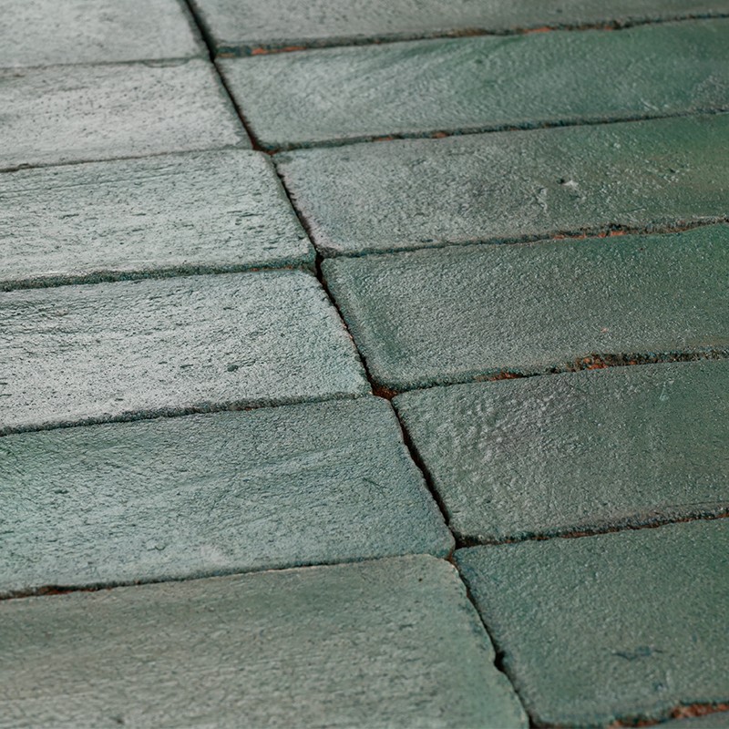 Azulejos verdes retangulares: piso com peças levemente desgastadas, juntas escuras, manchas acastanhadas e brilho irregular na superfície.
