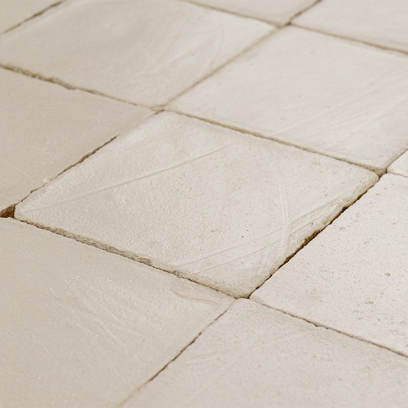 Azulejos cerâmicos quadrados bege, superfície texturizada com riscos finos; rejuntes irregulares entre peças, dispostos em padrão contínuo sob iluminação suave.