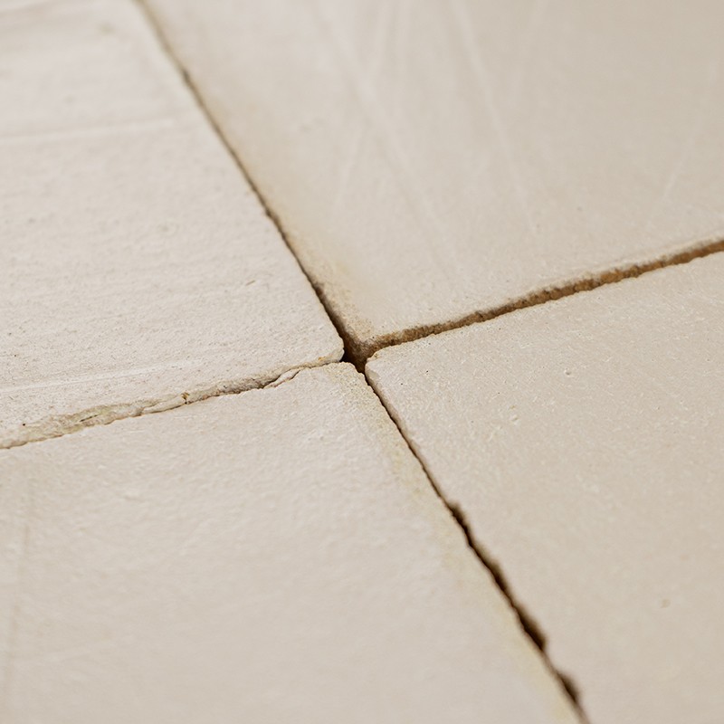 Vier matte beige Keramikfliesen mit schmalen Fugen und feinen Rissen; Nahaufnahme, leicht strukturierte Oberfläche, warmes Seitenlicht.