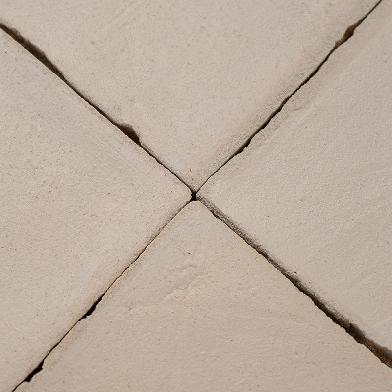Azulejo claro dividido en cuatro por juntas profundas; superficie rugosa con grietas finas y acumulación oscura en las uniones, rodeado por azulejos adyacentes.