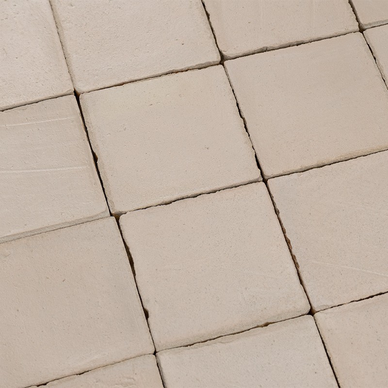 Azulejos cuadrados crema dispuestos en rejilla; juntas estrechas y oscuras, bordes desportillados y ligeras desalineaciones, superficie mate.