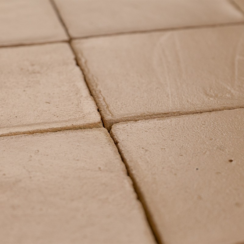 Beige keramische tegel met matte structuur; zichtbare voegen en lichte oppervlakte-onregelmatigheden, geplaatst naast vergelijkbare tegels in een nauw aansluitend patroon.