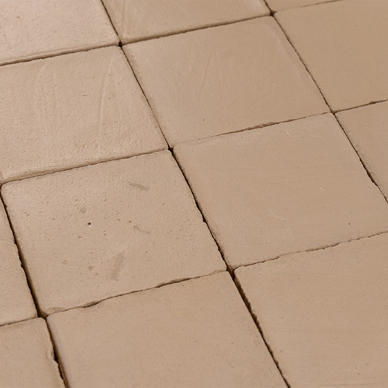 Vierkante beige keramische tegels met zichtbare voegen en lichte vlekken, gerangschikt in meerdere rijen op een vlakke vloer.
