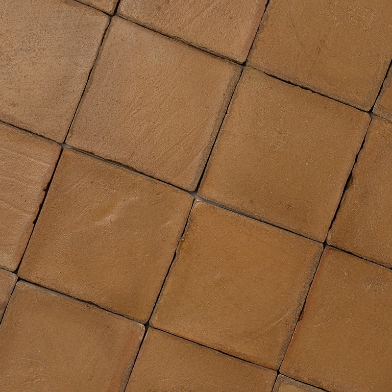 Azulejos quadrados cerâmicos terracota; rejunte escuro e irregular; superfície levemente desgastada; piso em padrão de grade, visto em close.