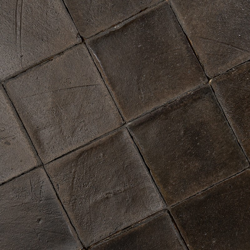 Azulejos quadrados escuros, superfície cerâmica irregular e gasta; alinhados em grade com finos rejuntes; aparência molhada ou suja, provavelmente piso interno ou área externa coberta.