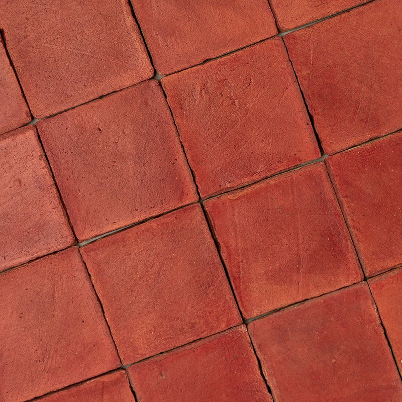 Worn red square tiles; dark joints forming a grid pattern, rough surfaces with stains and small misalignments, resembling outdoor flooring.