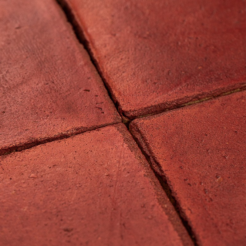 Azulejo rojo cuadrado con juntas pronunciadas formando una cruz; superficie ligeramente rugosa, con pequeños poros y desgaste leve, en un conjunto de pavimento o revestimiento cerámico.