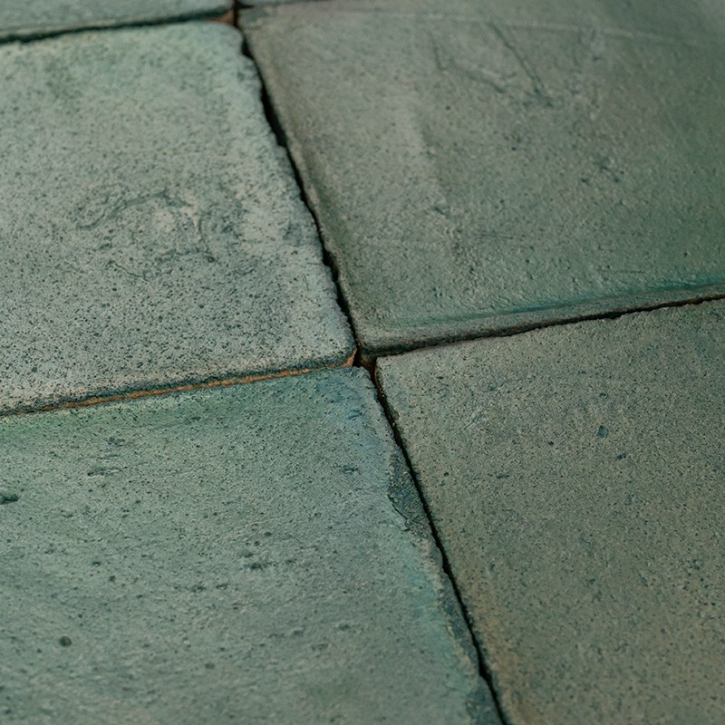 Blue-green ceramic tiles with textured surface and worn edges; dark joints forming a corner, exterior floor or patio appearance.
