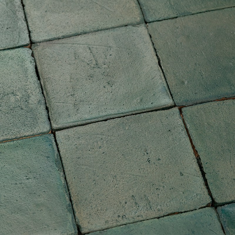 Azulejo quadrado verde-acinzentado com superfície levemente rugosa; juntas escuras visíveis, padrão repetido formando piso envelhecido.