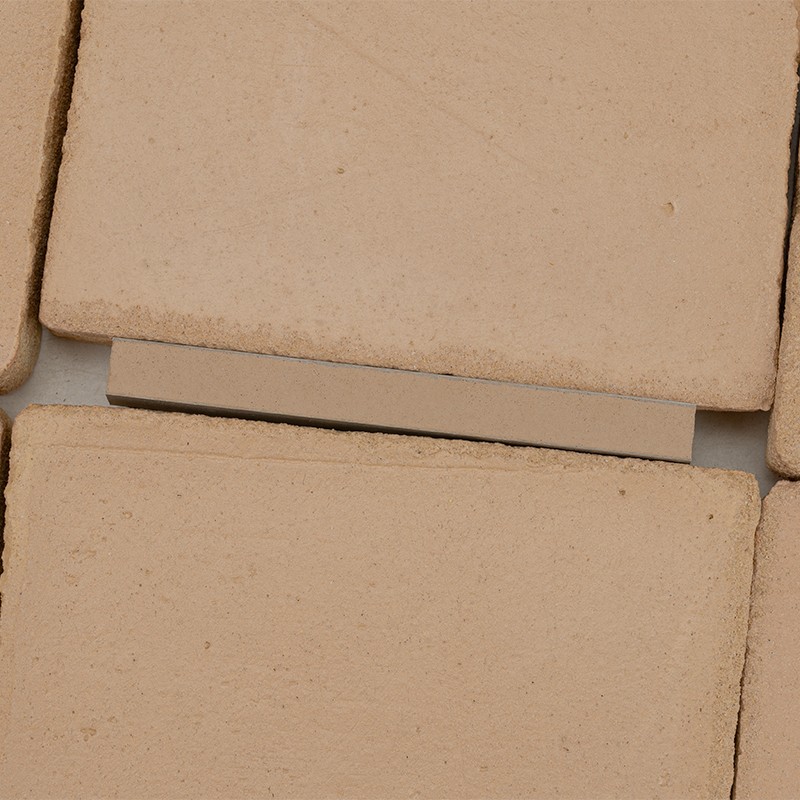 Beige keramische tegel met licht korrelige textuur; meerdere vierkante tegels liggen dicht tegen elkaar met donkere voegen en kleine kleurvariaties.