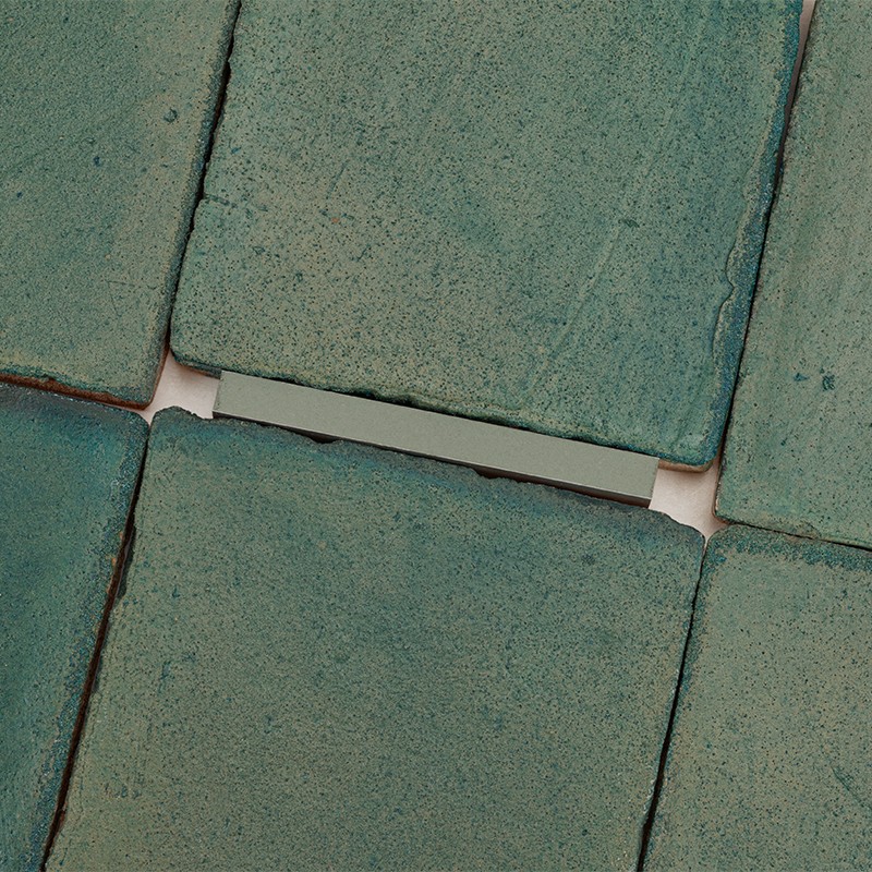 Azulejos quadrados verde-azulados texturizados; juntas largas e desiguais mostram espaços brancos entre peças levemente desalinhadas.