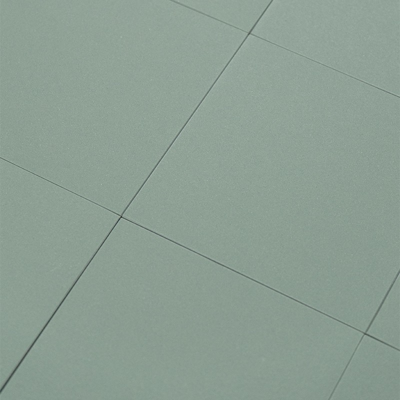 Azulejo verde mate como foco; juntas finas forman una cuadrícula y muestran varios azulejos contiguos dispuestos en ligero ángulo sobre una superficie lisa.