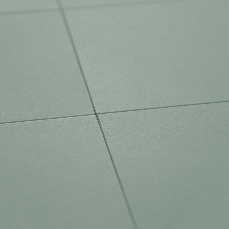 Glatt-matte hellgrün-graue Fliese, Kreuzung zweier schmaler, tiefer Fugen; dezente Schatten und feine Körnung, wahrscheinlich Teil eines Boden- oder Wandbelags.
