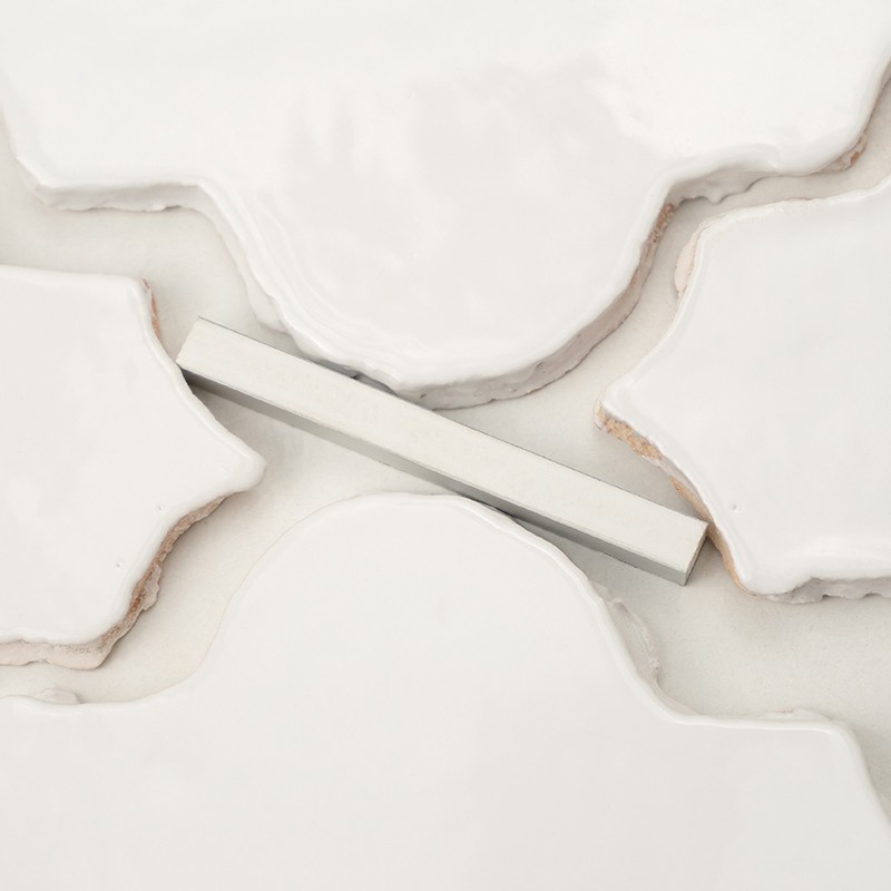 Weiße glasierte ornamentale Fliesen mit unregelmäßigen Konturen und sichtbaren Schnittkanten; eine längliche silberne Abstandshilfe liegt diagonal über den hellen Fugen.