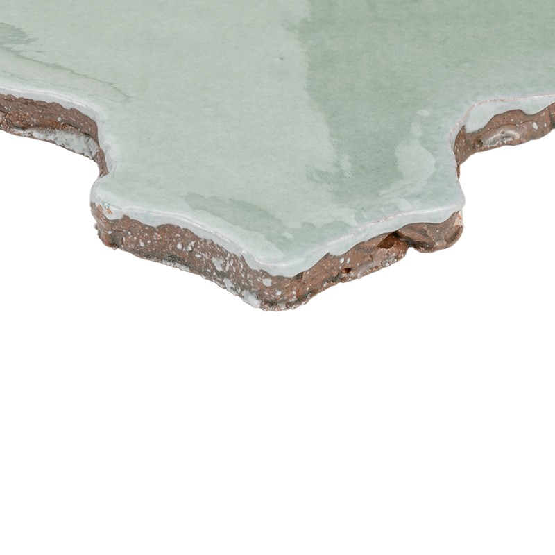 Azulejo esmaltado verde pálido con borde irregular y fracturado; base de arcilla marrón y capas de yeso blanco visibles, sobre fondo blanco.
