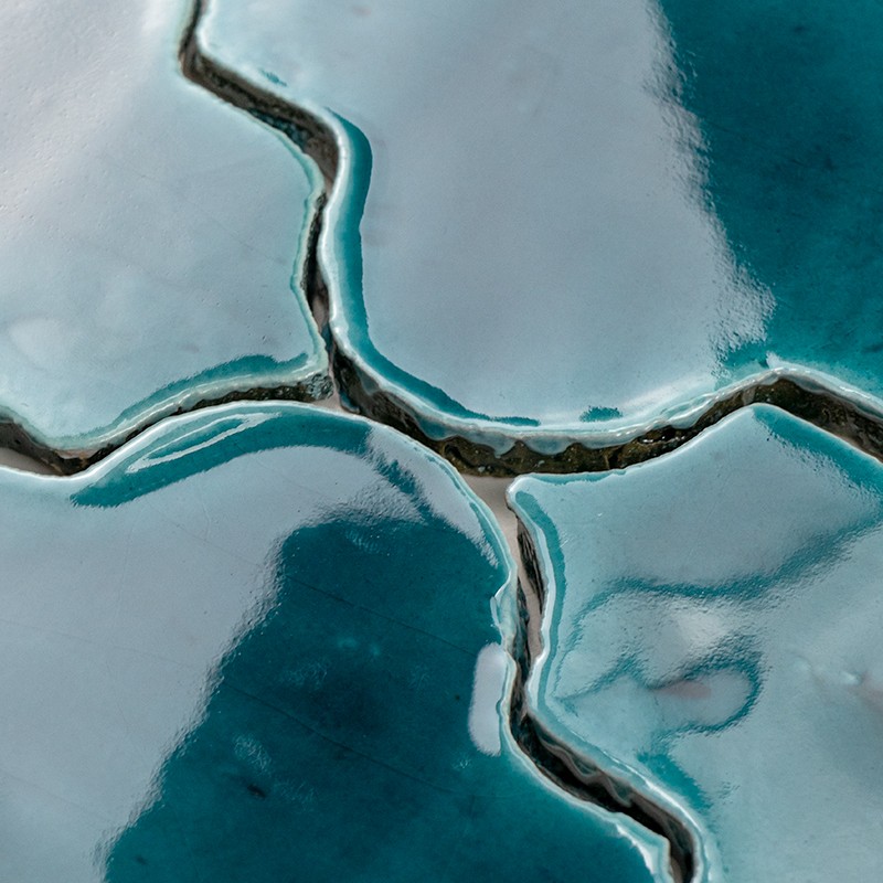 Carrelage vernissé turquoise et blanc, fissuré en croix; fissures sombres remplies d'eau formant canaux, surface brillante bordée de zones bleu profond.