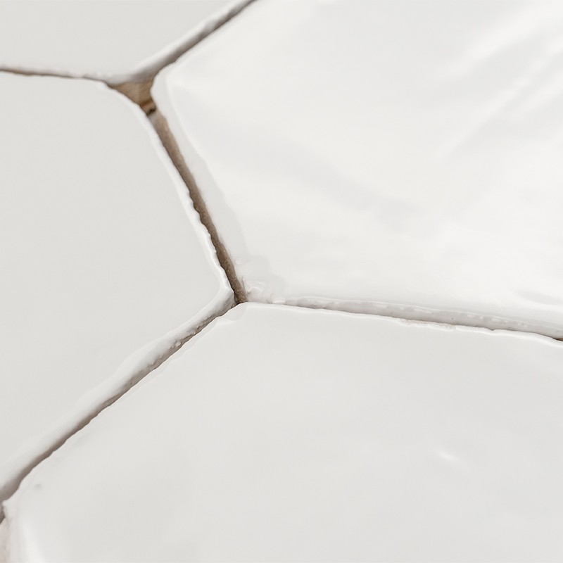 Carrelage hexagonal blanc en gros plan, joints irréguliers et légèrement salis, surfaces brillantes aux bords arrondis et fond neutre.