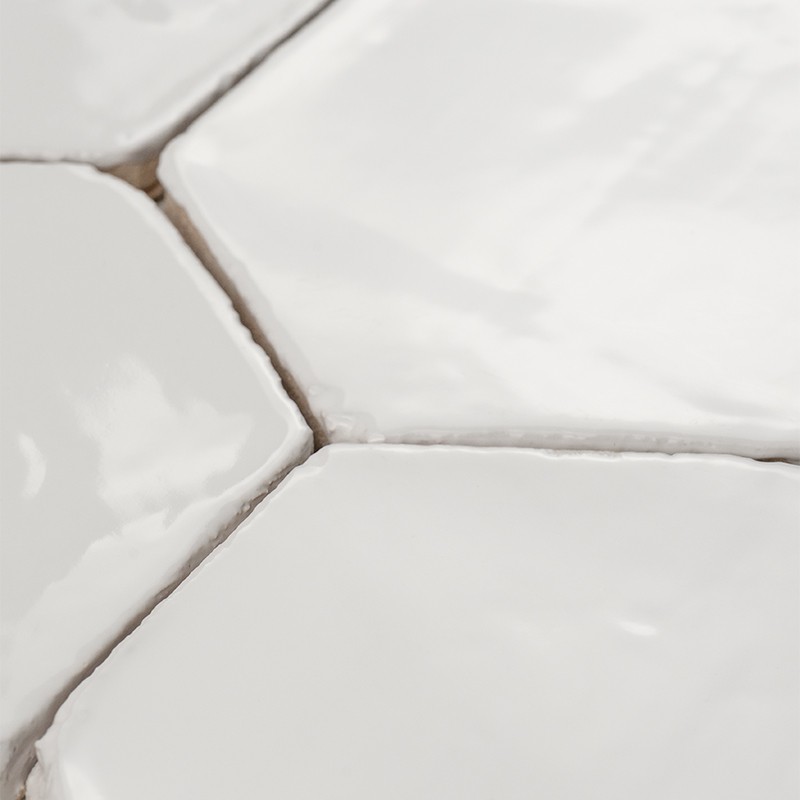 Azulejo blanco hexagonal brillante con superficie irregular y bordes curvos; lechada gris entre piezas y azulejos adyacentes formando un patrón continuo.