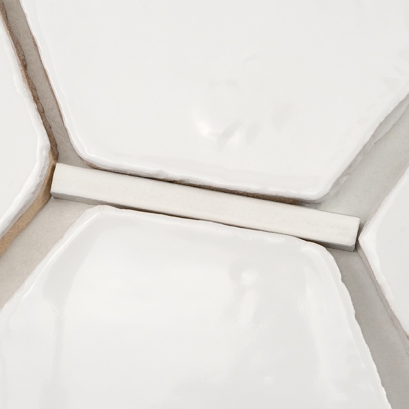 Azulejo cerámico blanco brillante, forma hexagonal con bordes redondeados; junta de mortero gris y espaciador rectangular blanco entre piezas; superficie vidriada con ligeras irregularidades.