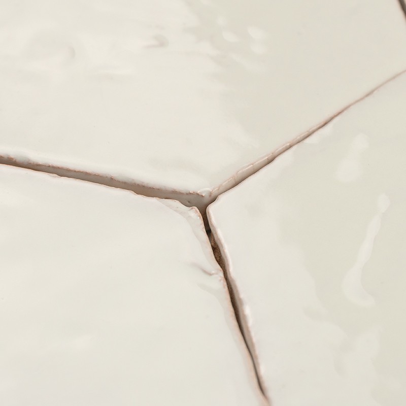 Azulejos blancos esmaltados (tres) con juntas en Y; lechada oscura y agrietada entre piezas, superficie brillante con reflejos y pequeñas imperfecciones.