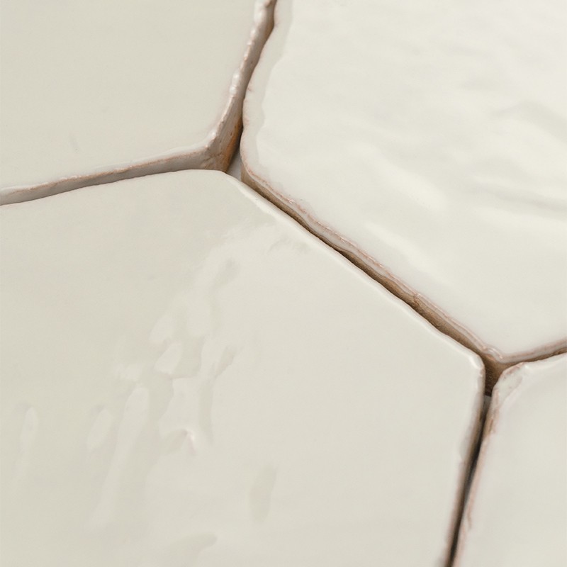 Carrelage hexagonal blanc brillant en gros plan, angle montrant la jonction de trois carreaux; joints beige clair légèrement irréguliers, surfaces lisses avec reflets.