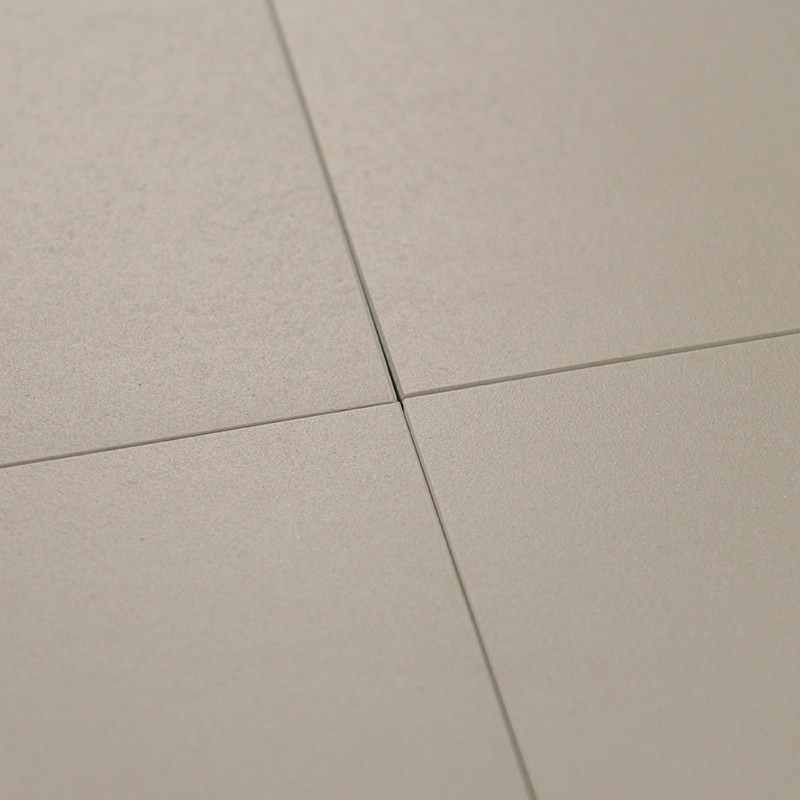 Azulejos quadrados bege fosco, quatro reunidos formando cruz de juntas finas escuras; superfície levemente texturizada, iluminação suave criando sombras discretas.