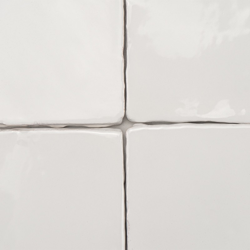 Cuatro azulejos cerámicos blancos brillantes, bordes redondeados; juntas grises en cruz, superficie ligeramente irregular y reflectante, encuadre en primer plano.