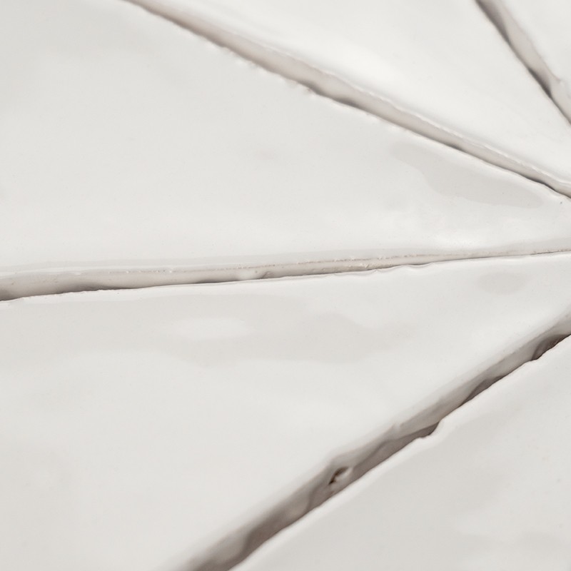 Azulejo cerámico blanco dividido por juntas profundas en forma radial, superficie vidriada con reflejos y sombras suaves; se aprecia unión en instalación cerámica cercana.