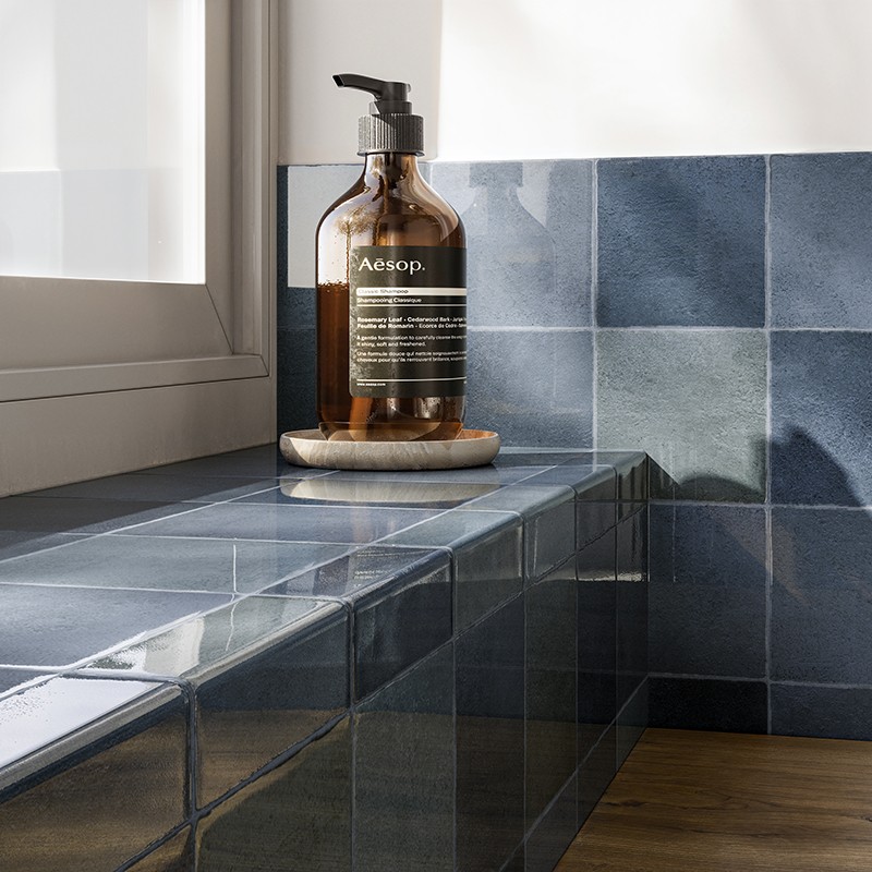 Azulejo cerámico azul brillante como foco, banco revestido junto a ventana, botella dispensadora ámbar de Aesop sobre posavasos, luz natural formando reflejos.  Texto visible: "Aesop." (el resto de la etiqueta no es legible).