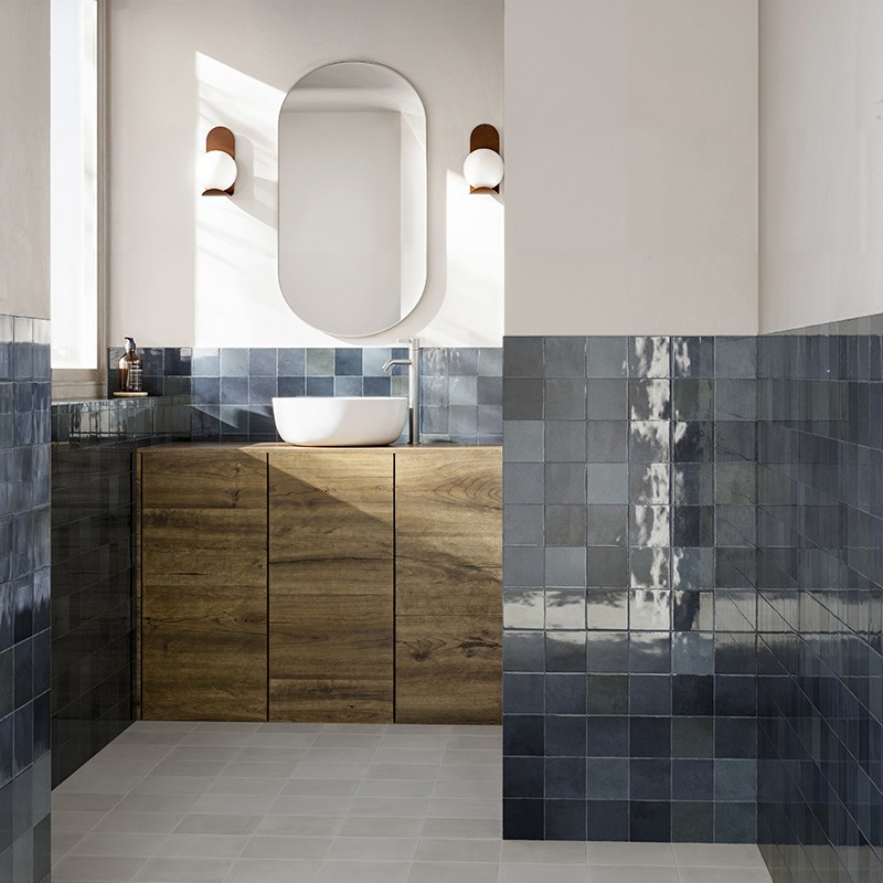 Azulejos cerámicos azul oscuro en semialzado; lavabo blanco de sobreponer sobre mueble de madera, espejo ovalado entre apliques y paredes blancas, suelo gris claro.