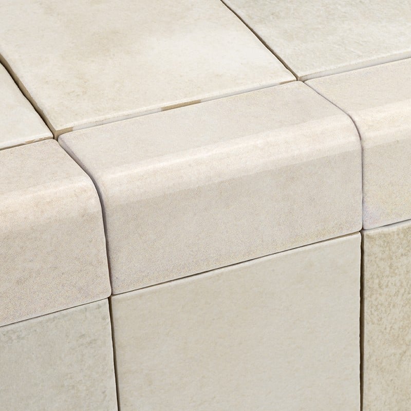 Azulejo cerámico blanco crema con borde redondeado; colocado en esquina de conjunto de azulejos similares, juntas finas visibles y textura mate ligeramente porosa.